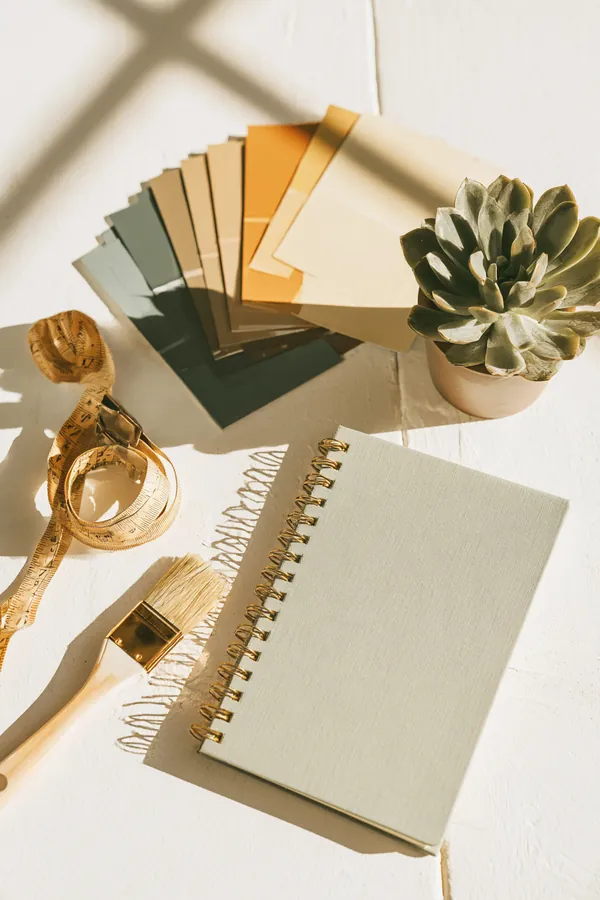 Flat lay of paint swatch cards, a measuring tape, a small potted succulent and a modern notebook on a white table