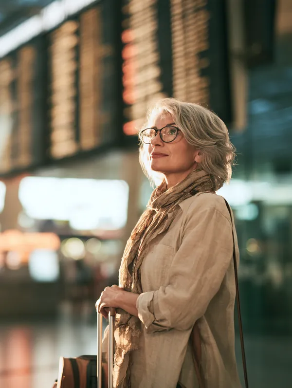 A woman in her 60s with a carry-on suitcase checks the departure board in a bright airport terminal