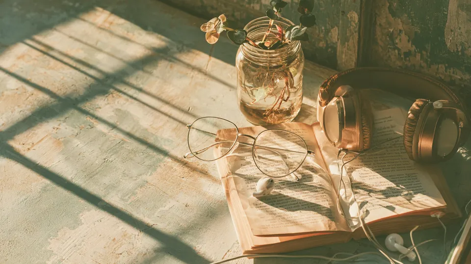Reading glasses, an open paperback, wireless headphones and a plant cutting in a glass jar on a pale wood table, set up for reading the Tripoint Lending blog