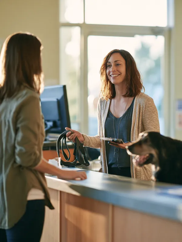 Woman in her 40s talking with a receptionist at a veterinary clinic desk about payment options while holding her dog's leash