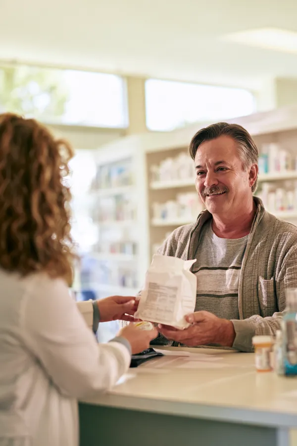 Pharmacist handing a prescription bag to an older man at a pharmacy counter, the kind of everyday cost covered in Tripoint Lending FAQs