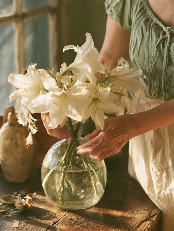 An older woman's hands arranging white lilies in a glass vase on a sideboard in soft window light